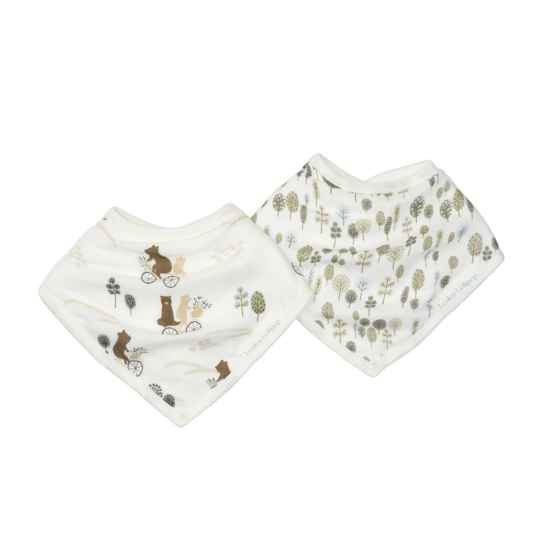 Two baby bandana bibs in a warm off-white color, one featuring a brown woodland squirrel and bicycle print, and the other featuring a green and brown forest tree print. Both are laid flat on a light gray background.