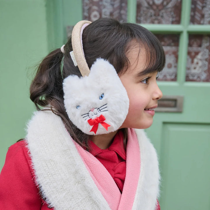 Child wearing white cat-themed earmuffs with a red bow in front of a green door.