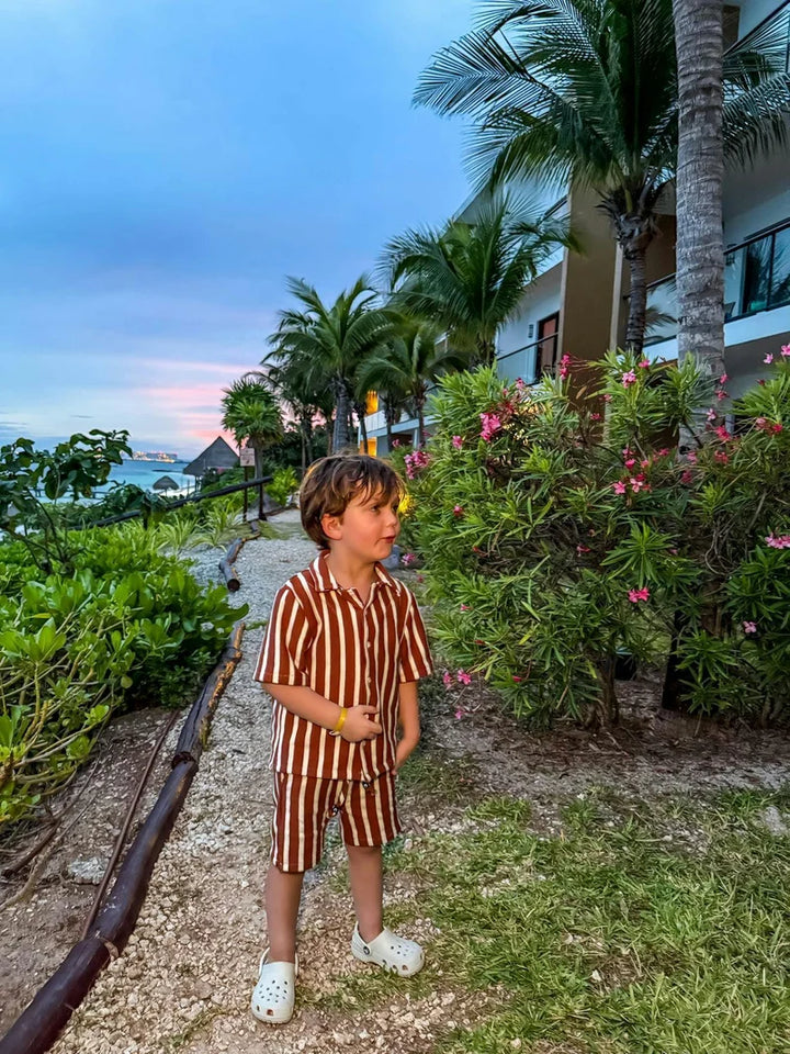 Child in a striped outfit standing in a tropical setting with palm trees and flowers.