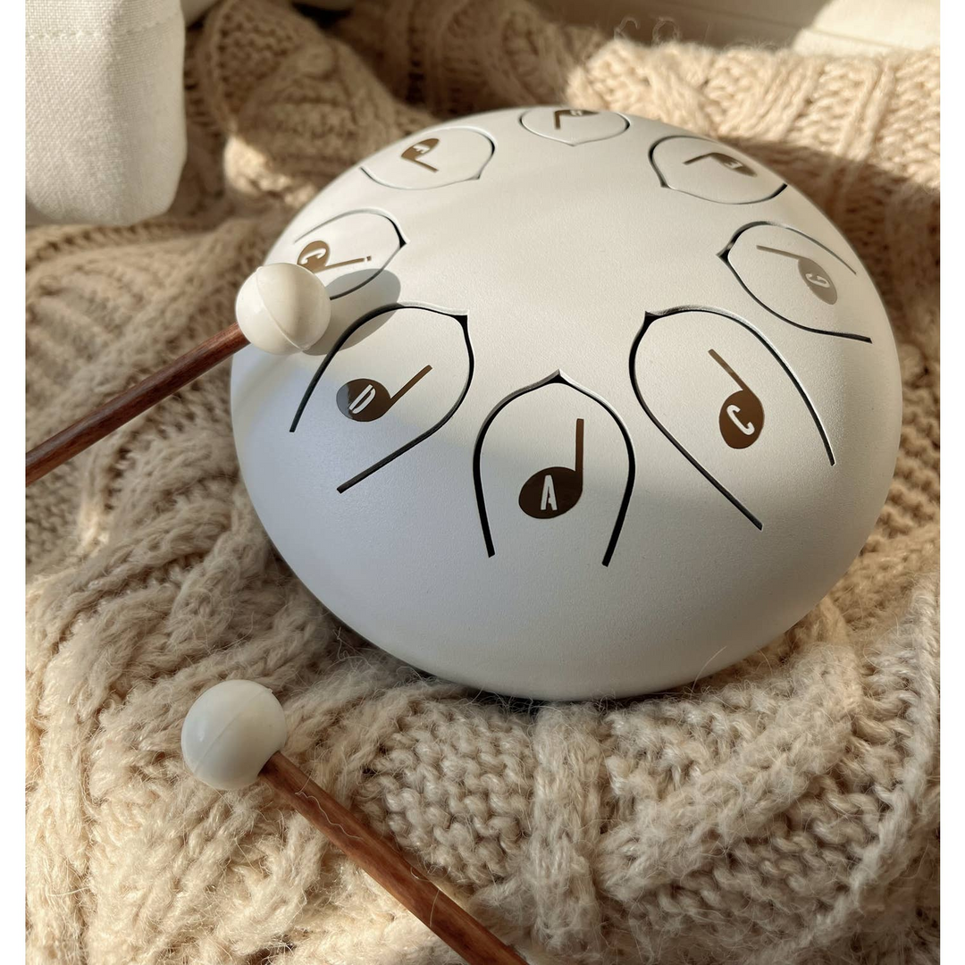 A white steel pan handpan with engraved musical note symbols, resting on a beige chunky knit blanket with two wooden mallets with white tips.