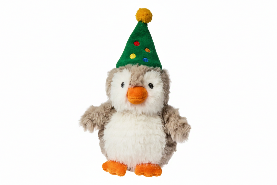 A plush toy penguin with fluffy white and gray fur, an orange beak and feet, wearing a green party hat with colorful dots and a yellow pom-pom, isolated on a white background.